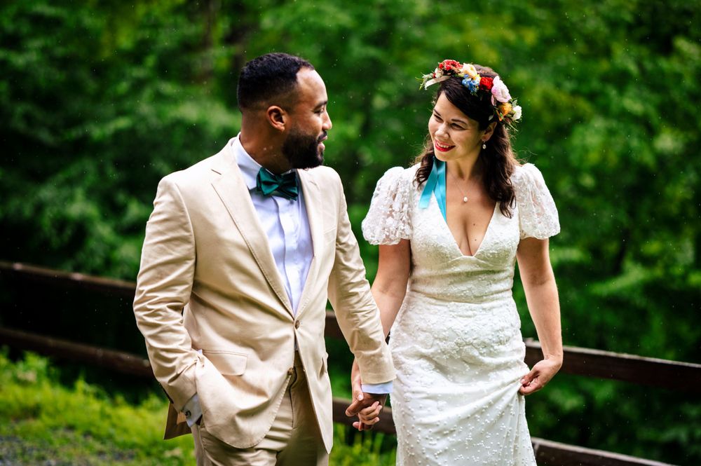Newlyweds holding hands and smiling at each other, groom in tan suit with colorful bow tie outdoors.