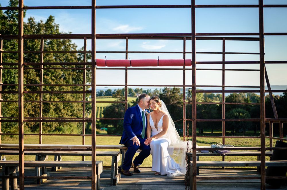 Bride and groom share a romantic moment sitting together within a rustic metal frame structure with green fields behind.