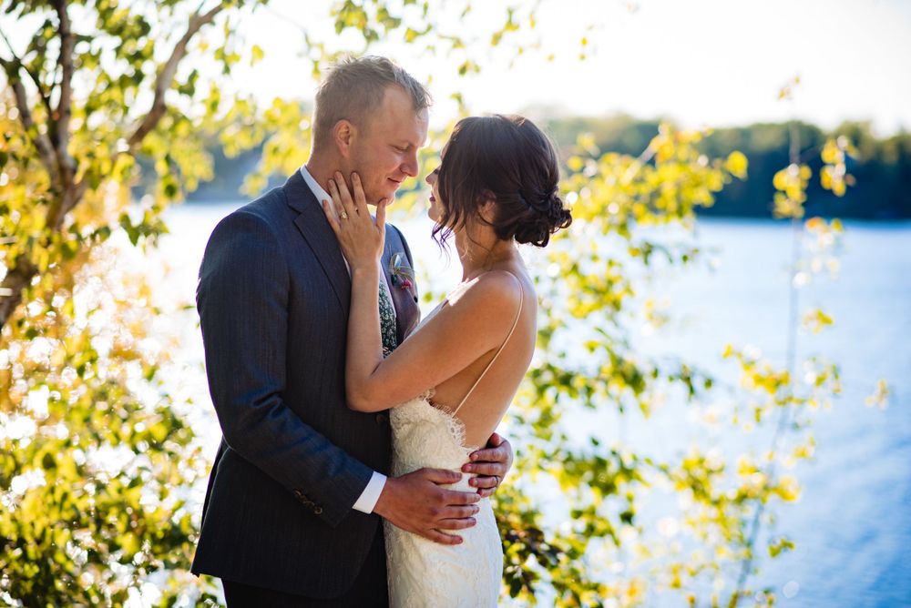 Bride and groom share a tender embrace near a sunlit lake surrounded by golden leaves on their wedding day.
