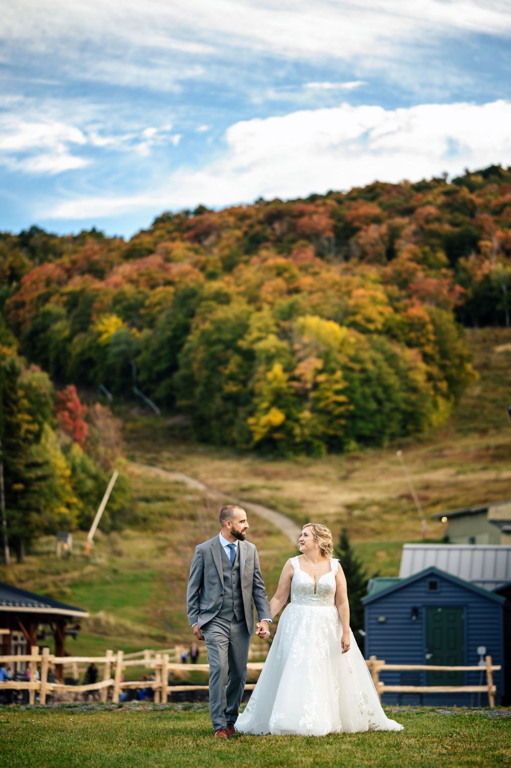 Bride and groom walk hand in hand on a hillside with vibrant autumn foliage covering the mountain behind them.