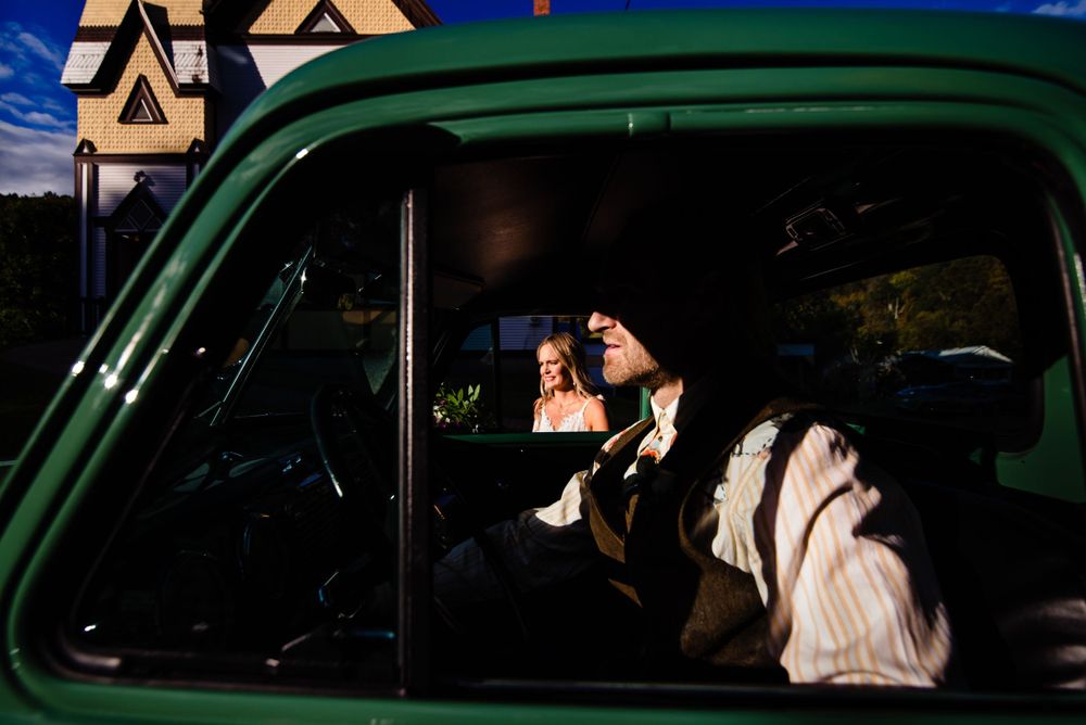 Bride and groom share romantic moment reflected in window of vintage green car at sunset.