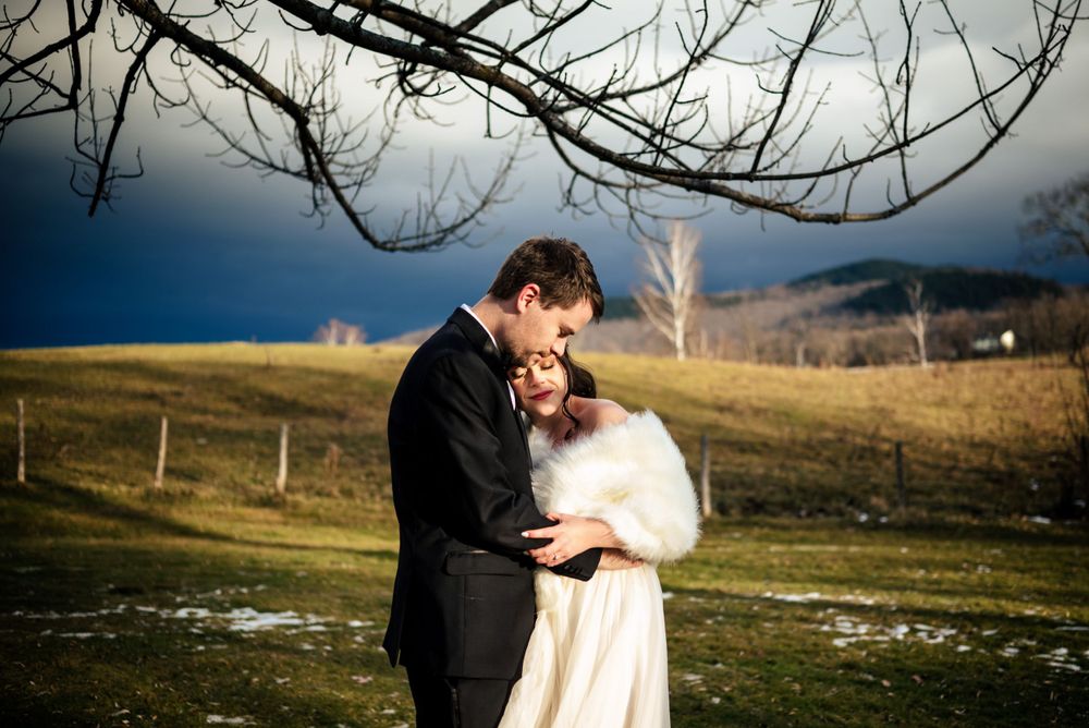 Bride and groom embrace outdoors in winter landscape with dramatic stormy sky and rolling green hills.
