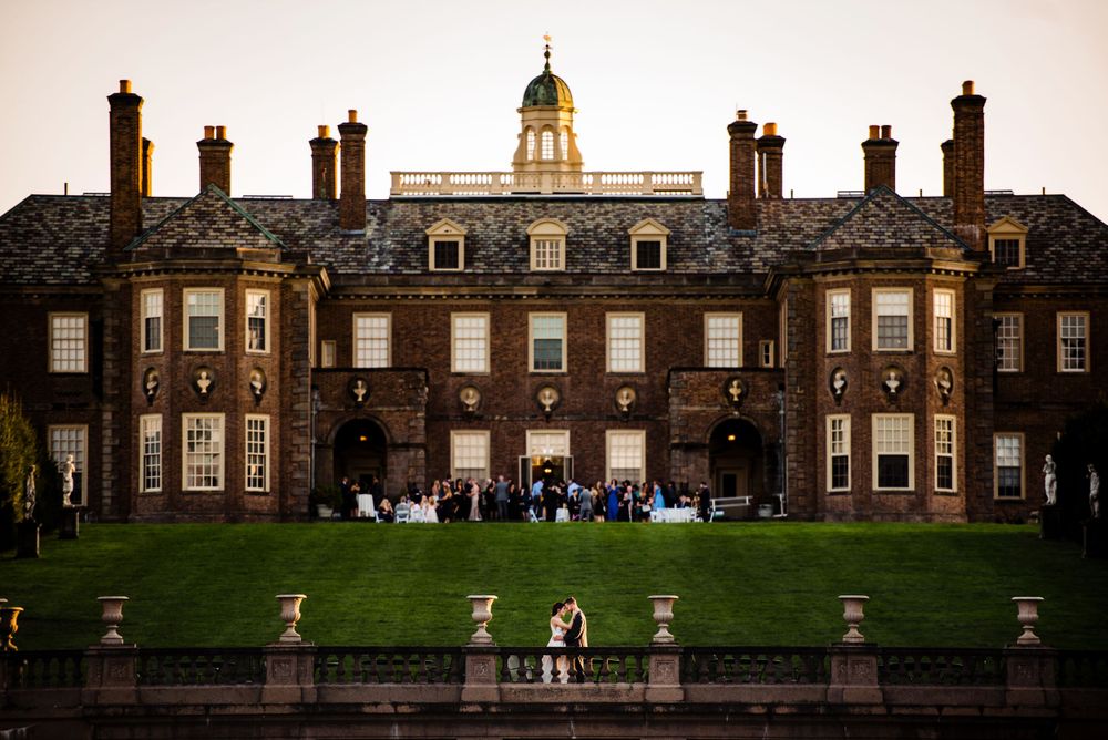 Wedding guests gather on lawn of grand historic brick mansion with domed cupola at dusk.