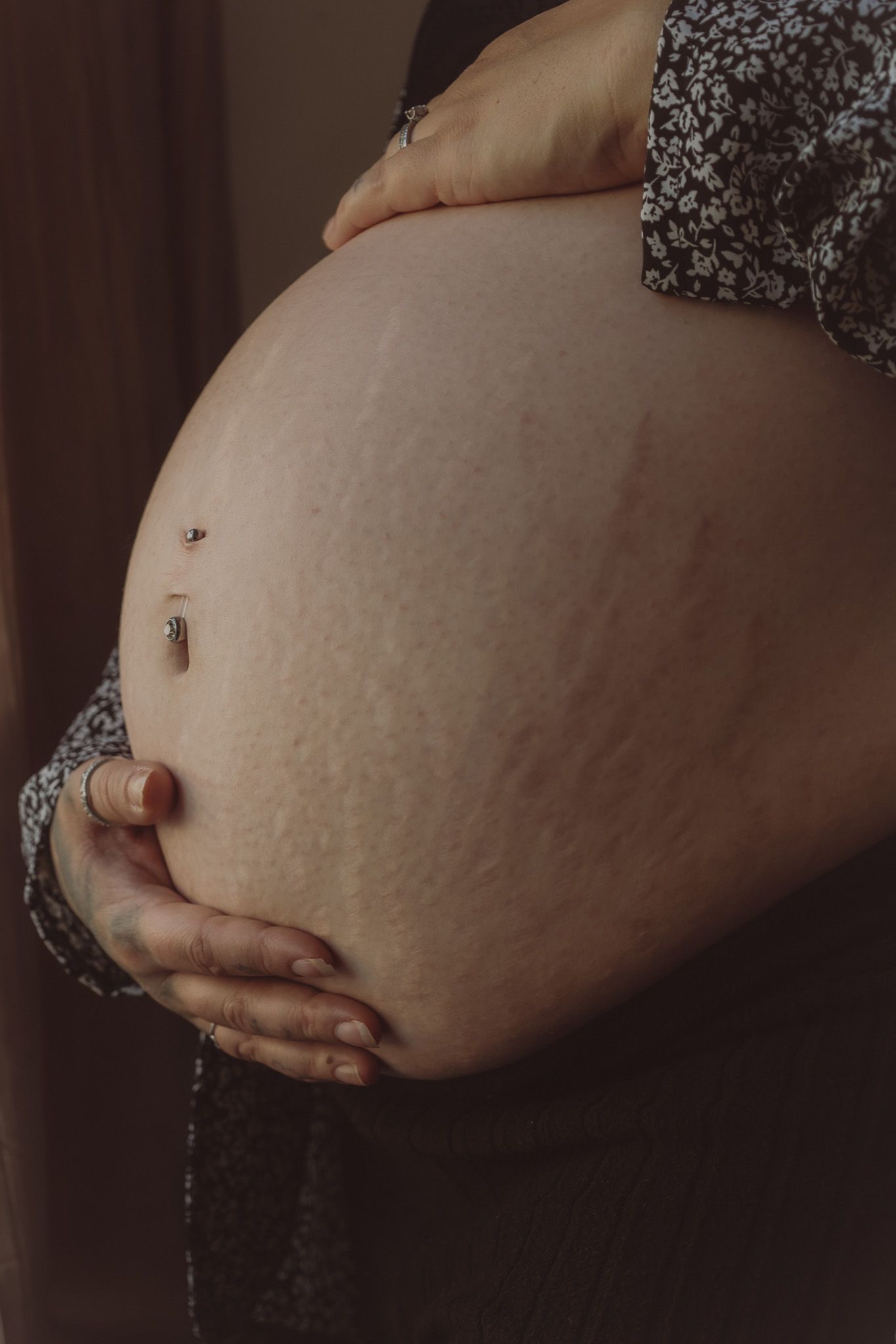 Close up of side view of pregnant belly, warm ,moody tone, belly button pierced, hands holding belly, maternity photography in suffolk