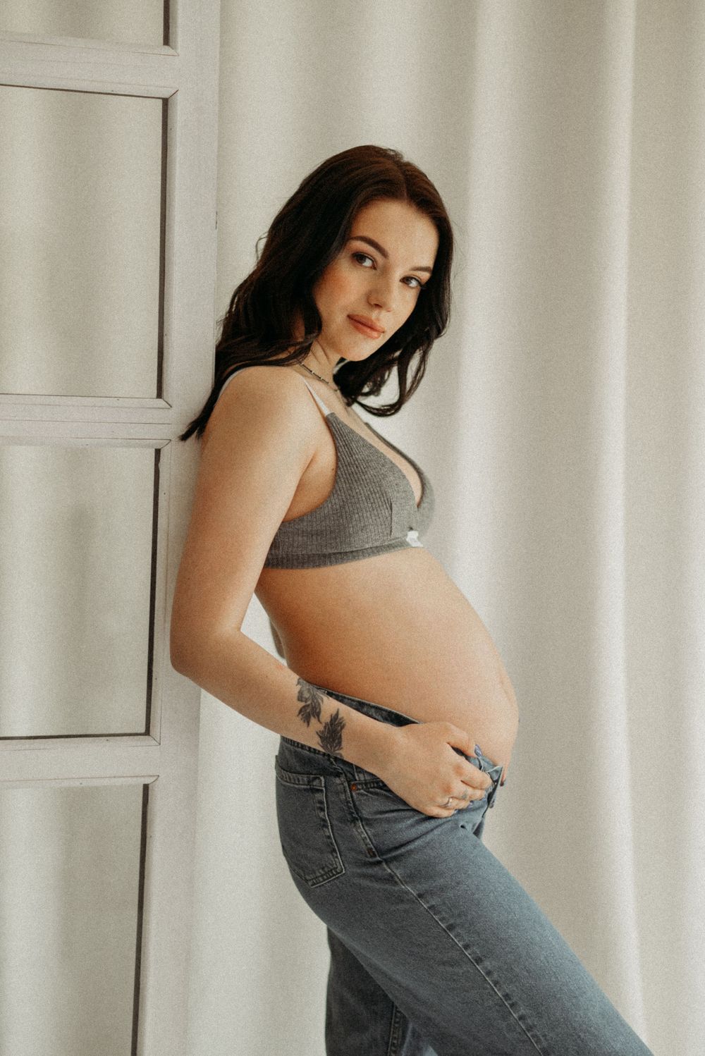 Casual maternity portrait in gray crop top and jeans against white wall.