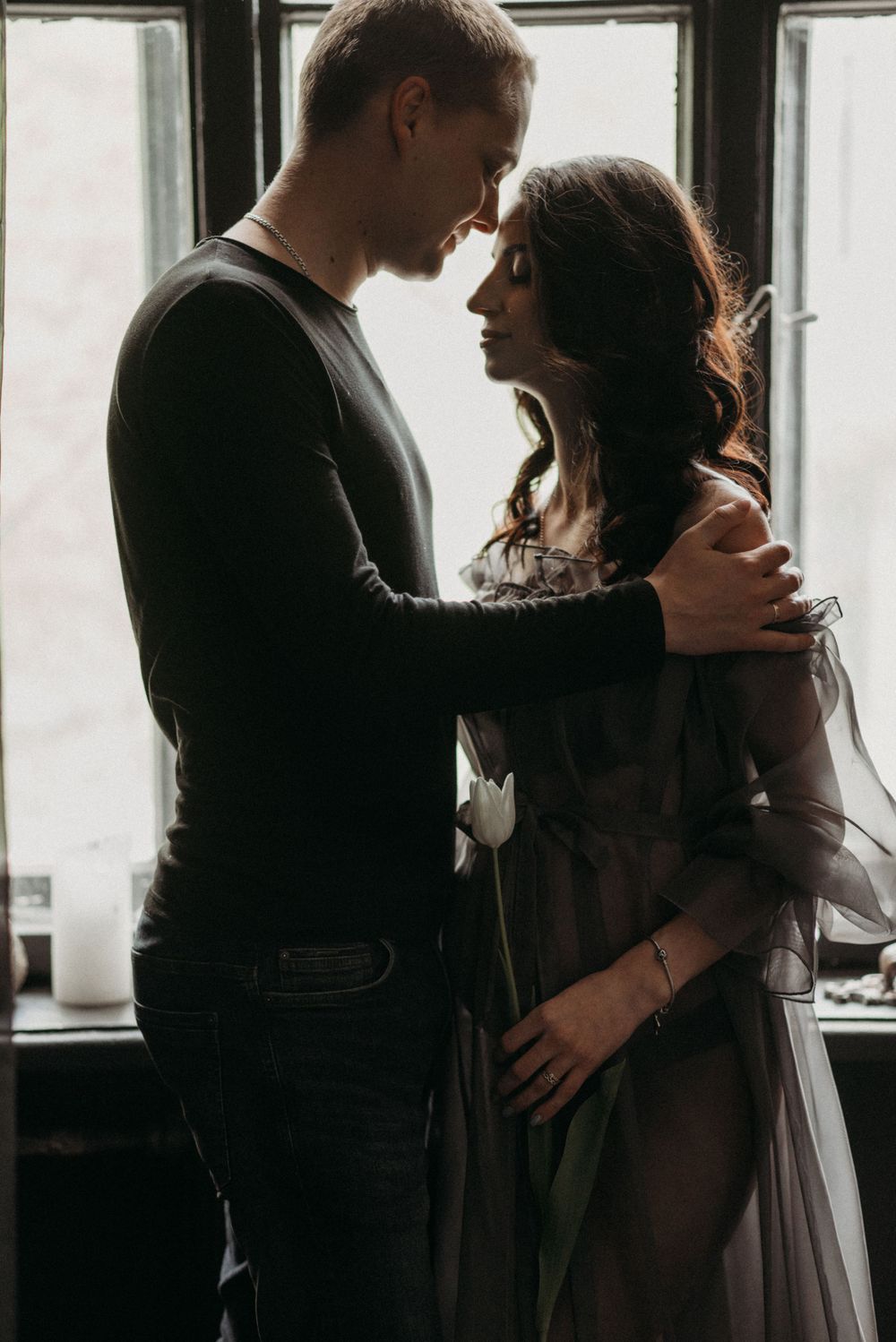 Romantic couple embracing by window in moody lighting with vintage aesthetic.