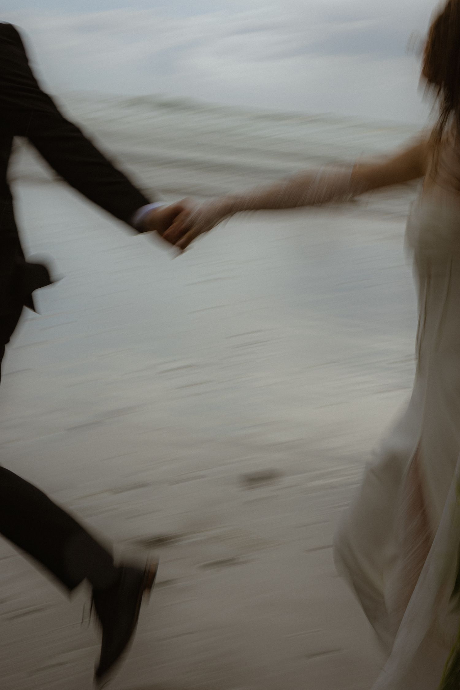 Blurred motion photo of a bride and groom holding hands while running on a beach in wedding attire.