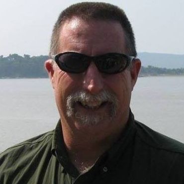 Man in green shirt and sunglasses smiles outdoors with water in background.