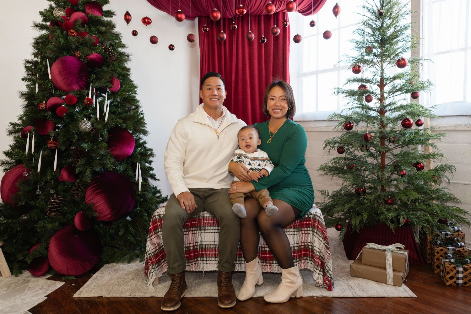Holiday family photo with Christmas trees, red decorations, and plaid accents in a bright festive setting.