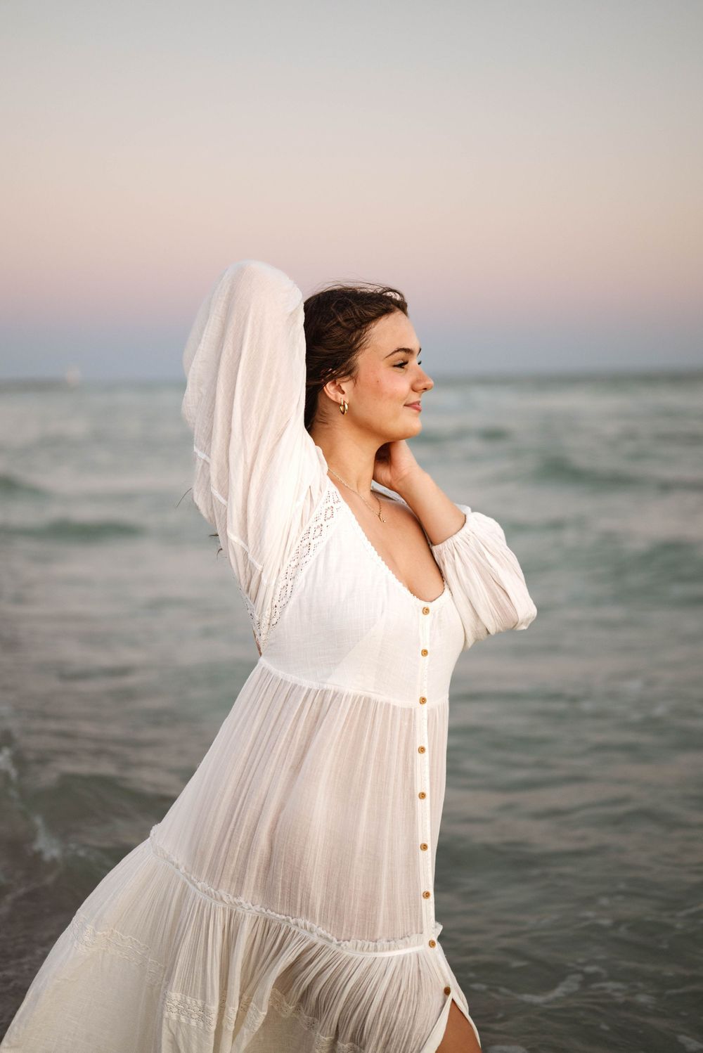 Woman in flowing white dress poses against ocean backdrop during golden hour.