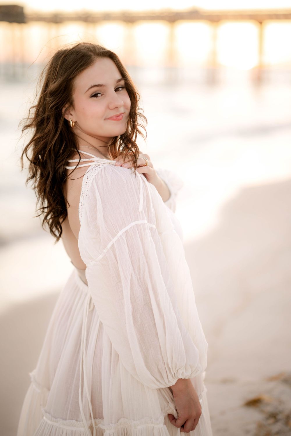 Ethereal beach portrait featuring a flowing white dress and romantic sunset lighting through ocean pier.