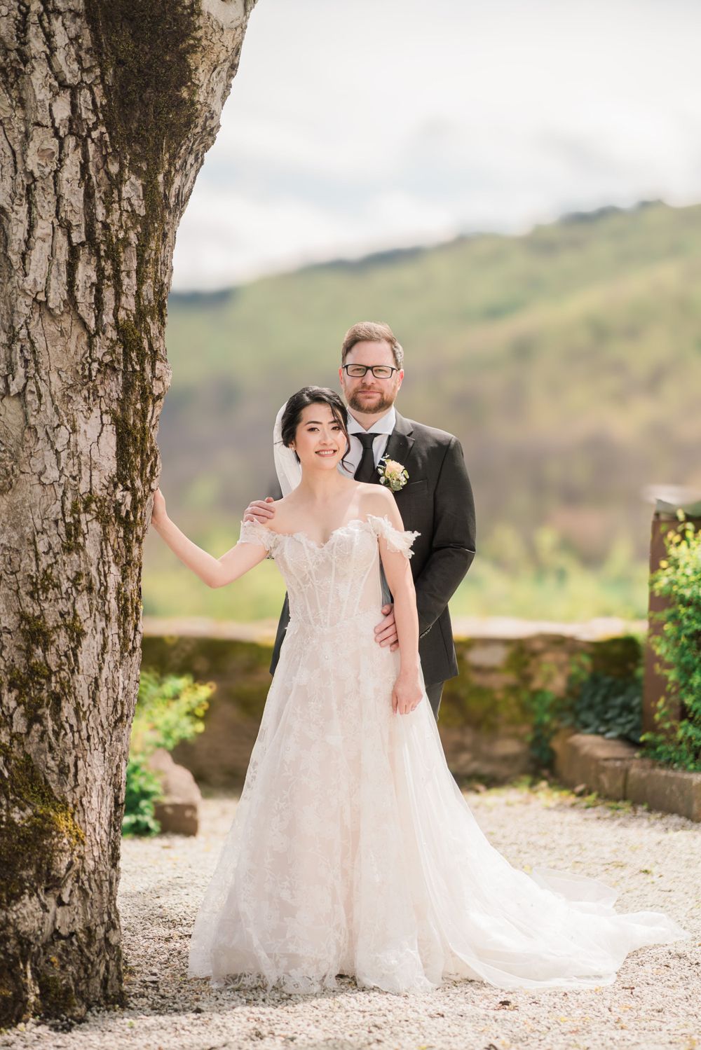 Wedding couple poses elegantly against large tree trunk in scenic mountain setting.