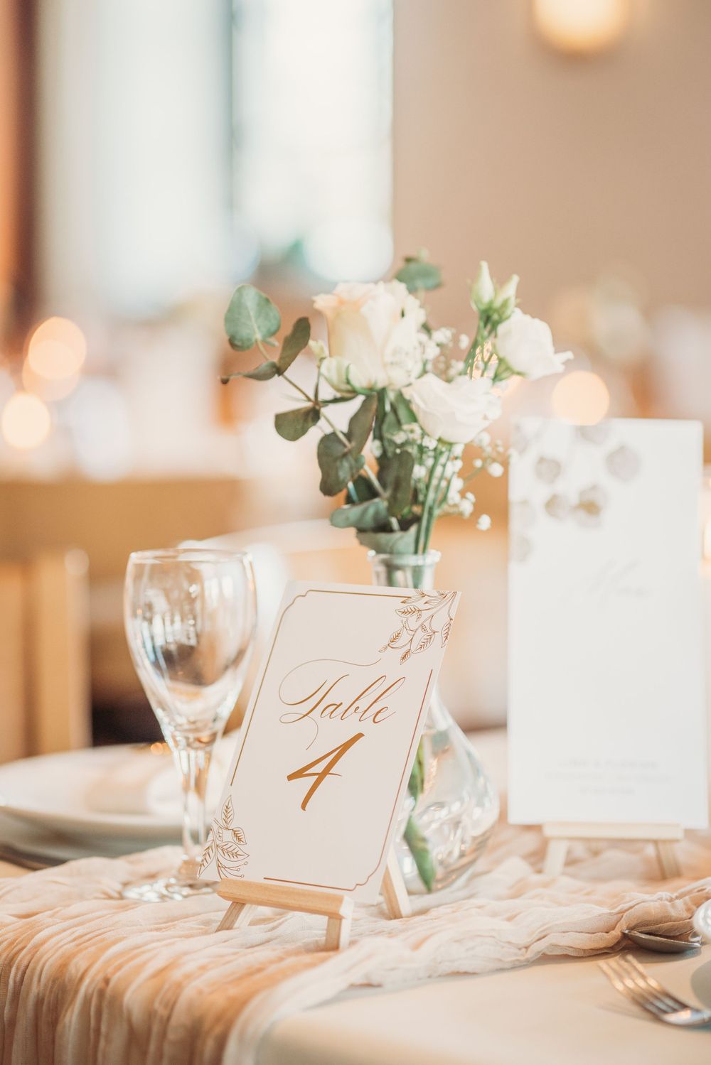 Elegant wedding table setting with table number 4, white flowers, and wine glasses on a cream tablecloth.
