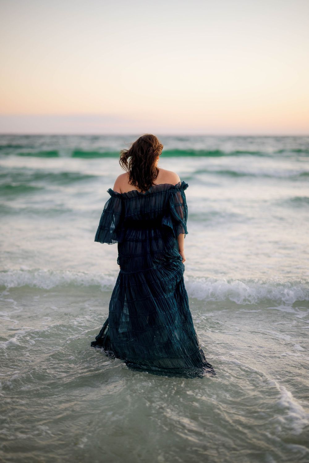 Back view of flowing navy dress against sunset waves on serene beach.