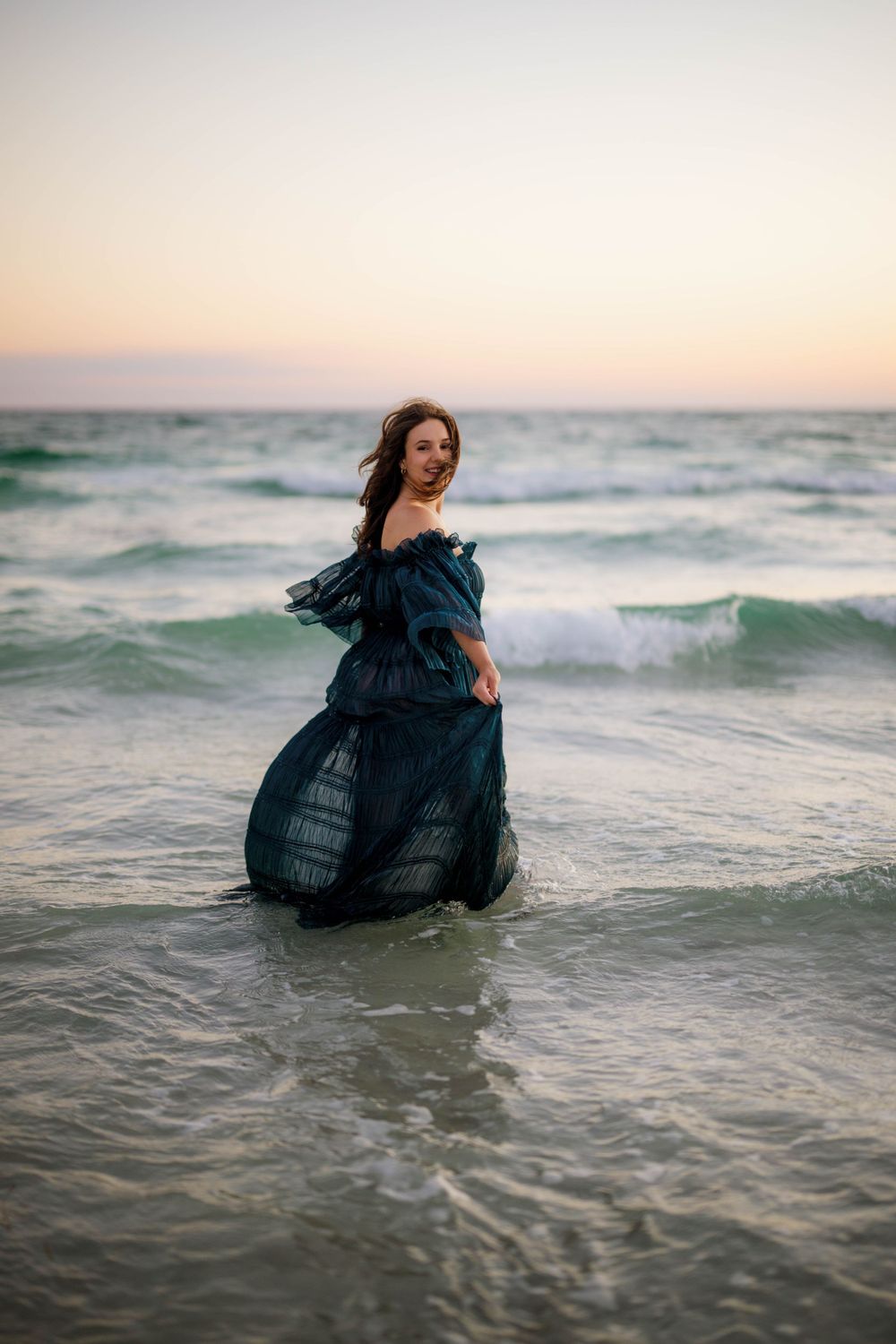Expectant mother in navy dress standing in ocean waves during golden hour.