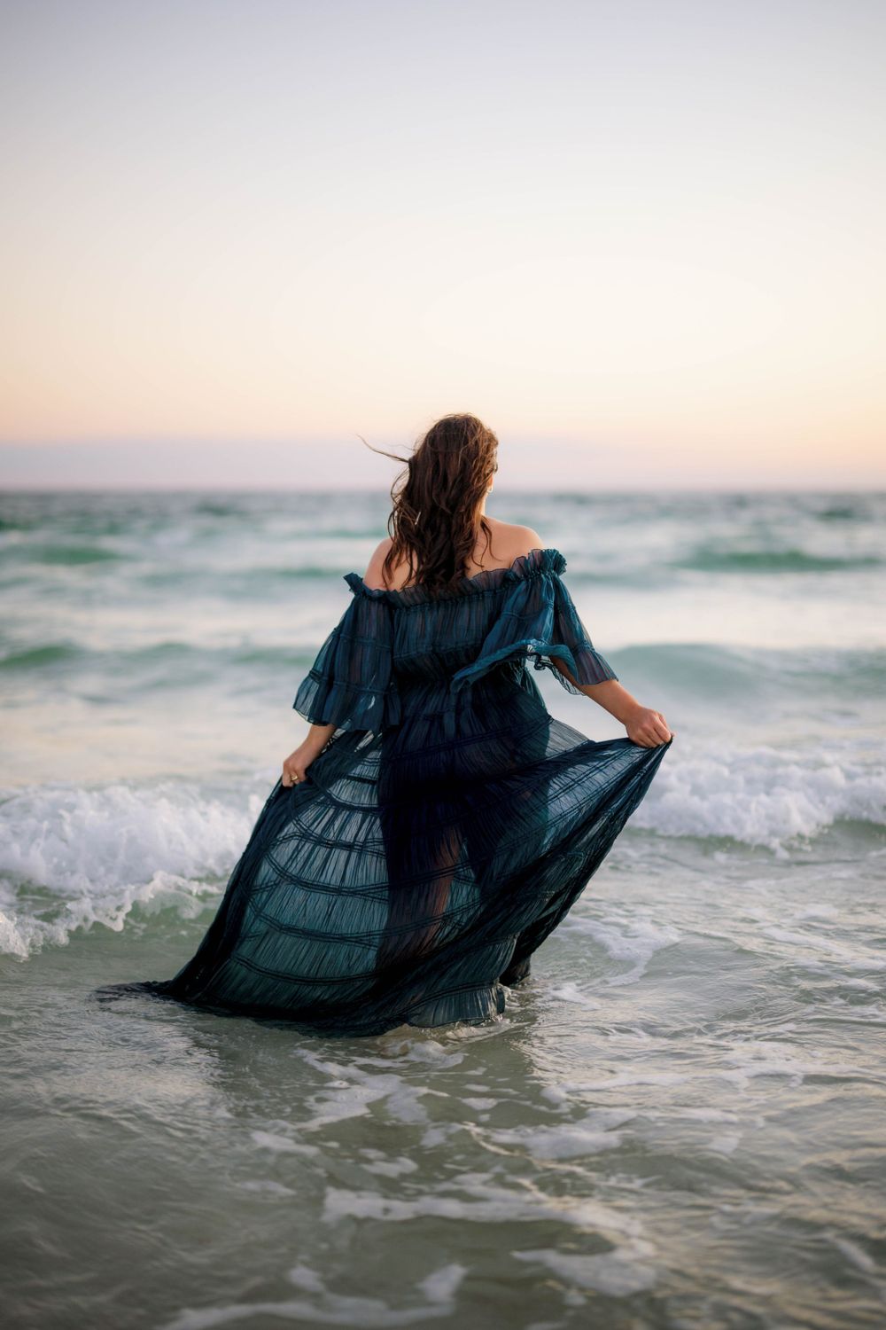 Flowing navy dress catches sea breeze during beach maternity photoshoot at dusk.