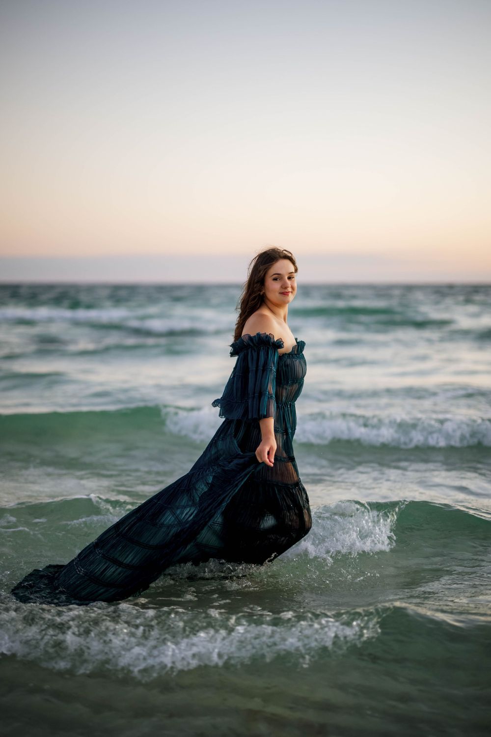 Dramatic beach maternity photo with navy dress flowing in ocean waves at sunset.