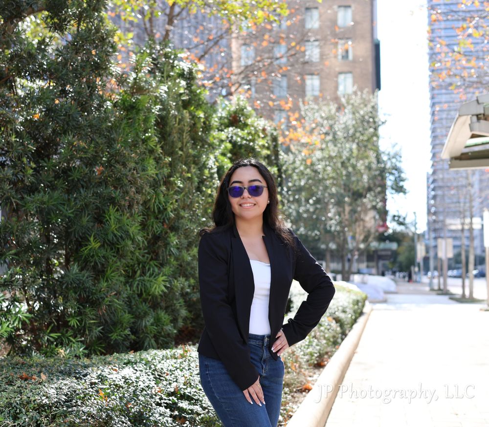 Woman in business attire poses outdoors near urban buildings and trees.