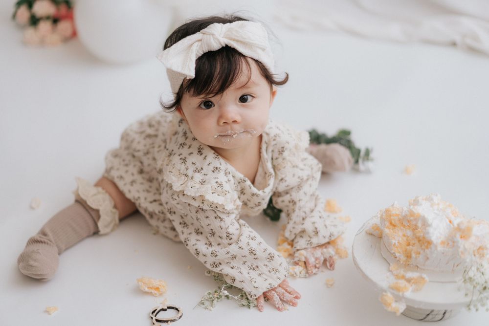 Baby in white lace dress crawls on floor near scattered orange flower petals during cake smash photo session.