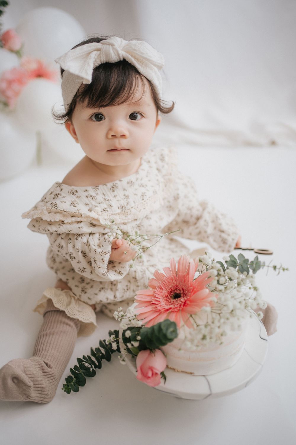 A baby in a white lace dress sits on the floor with a pink gerbera daisy flower and delicate baby's breath blooms.