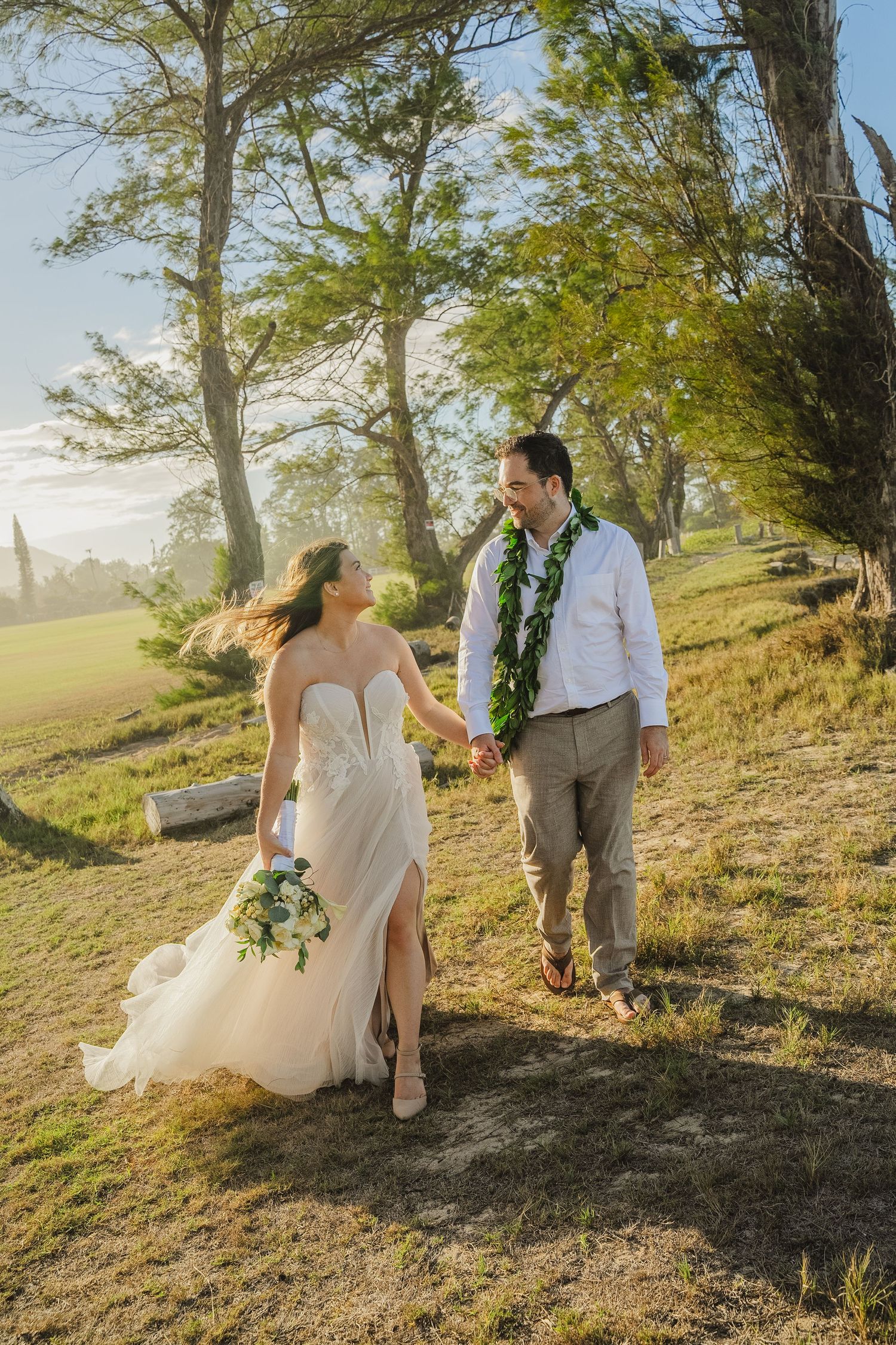 A couple in wedding attire sits together on a grassy hillside during golden hour with trees in the background.