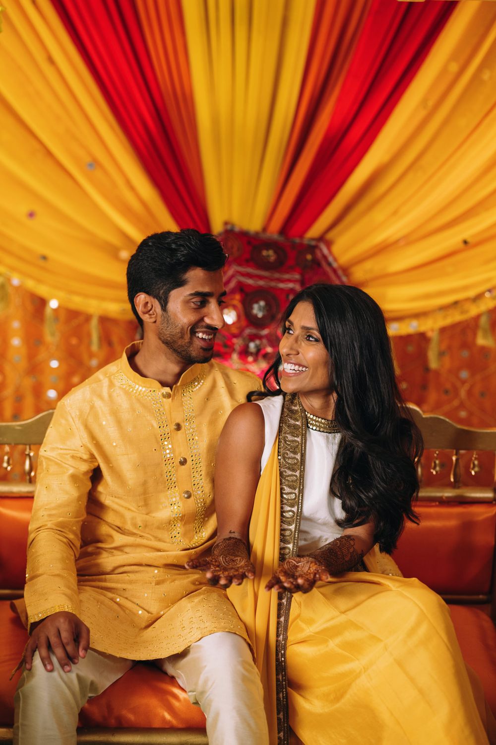 A couple in traditional yellow Indian attire share romantic moments on an ornately decorated stage with red and gold drapes.