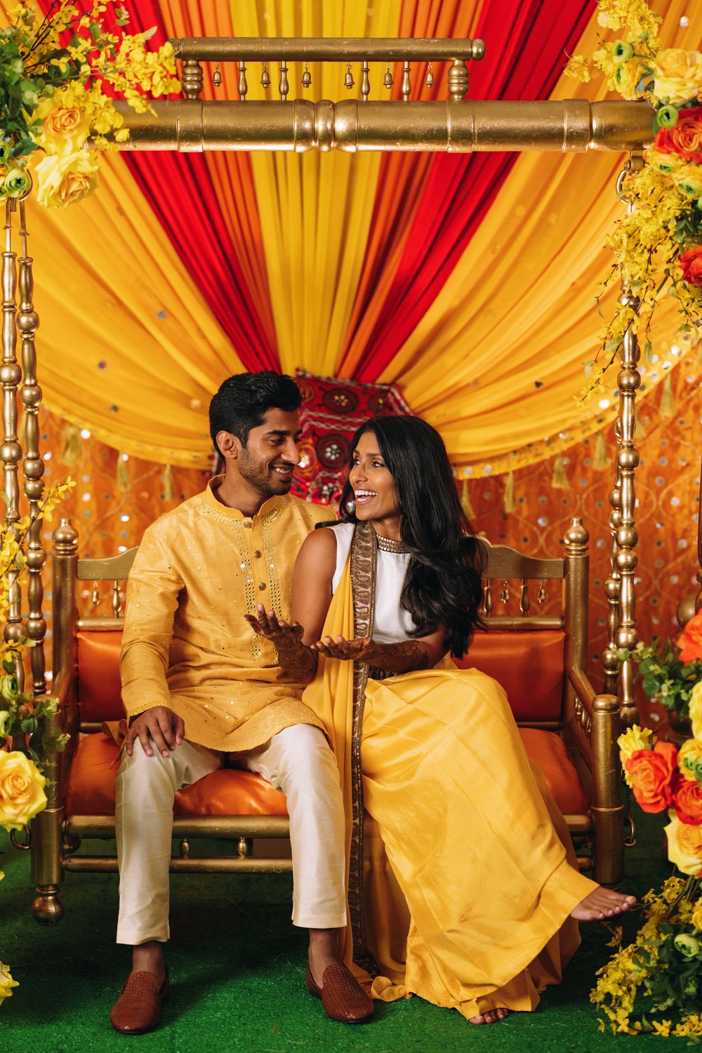 A romantic couple in matching yellow outfits poses together against a vibrant red and gold draped backdrop with floral decor.