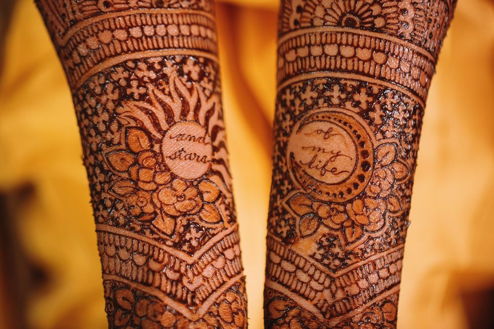 Intricate sun and moon henna mehndi design adorns arms with detailed patterns against warm golden background.