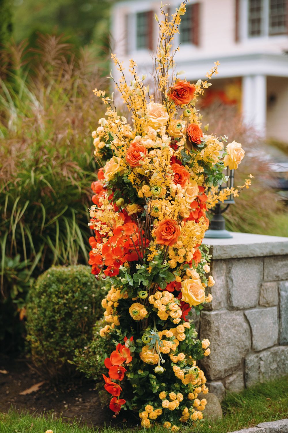 Tall floral arrangement with orange and yellow blooms cascading down a stone wall in a garden setting.