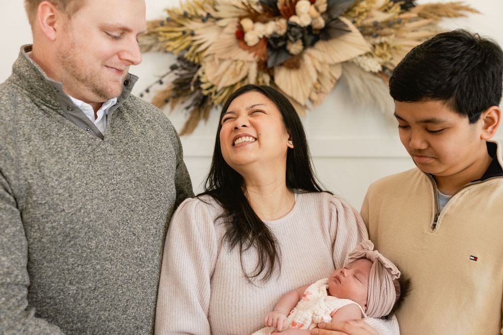 Family gathers around newborn baby in front of decorative dried floral arrangement.
