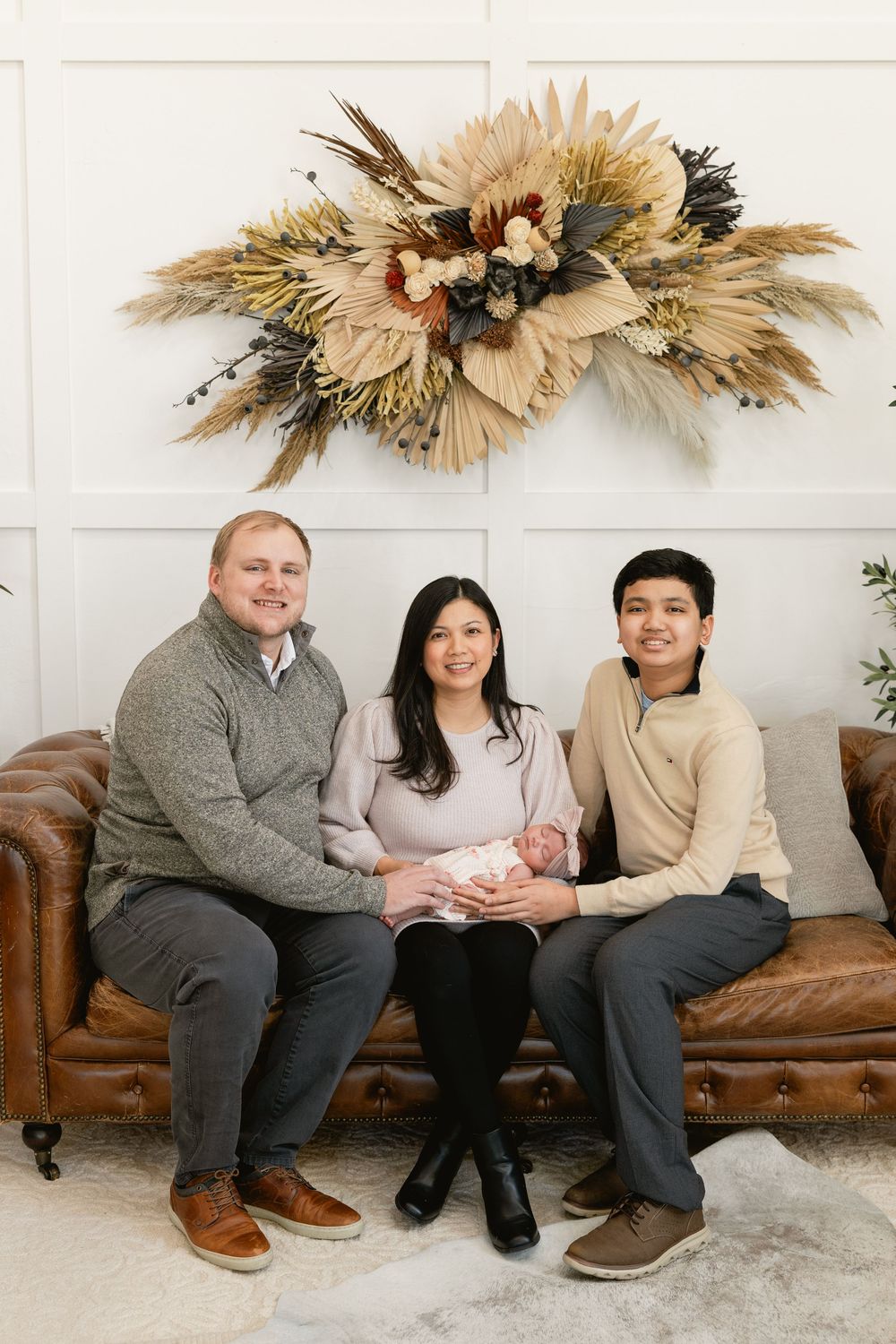 Family sits on brown leather couch with dried floral wall decor above.