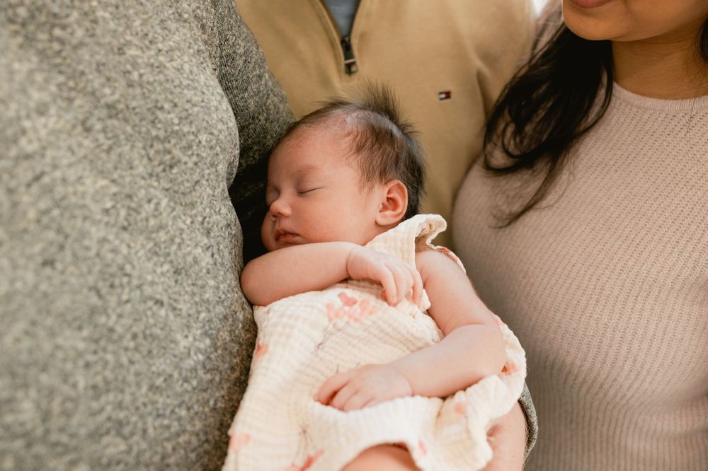 A newborn baby wearing a light colored onesie sleeps peacefully on a soft surface.