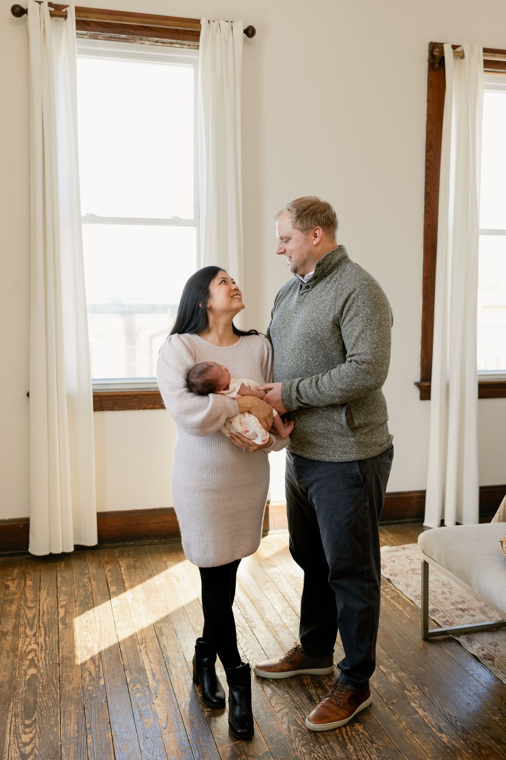 A couple dances together on hardwood floors in front of white curtained windows.