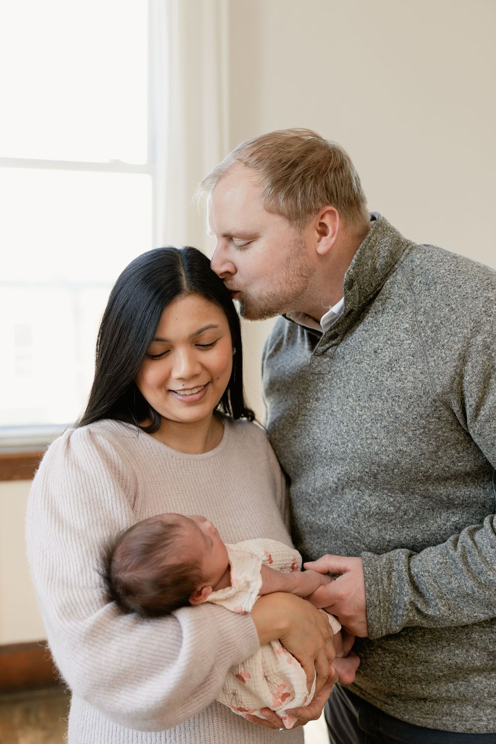 A couple in cozy sweaters share tender moments while holding their newborn baby in a bright room.