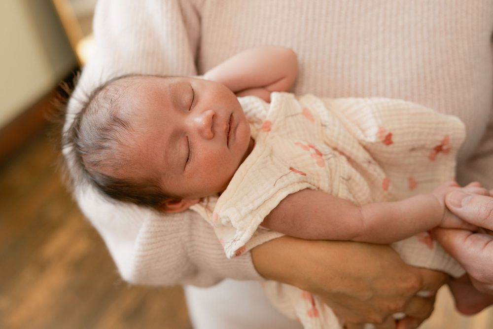 Parents cradle their sleeping newborn baby while wearing neutral colored sweaters in a sunlit room.