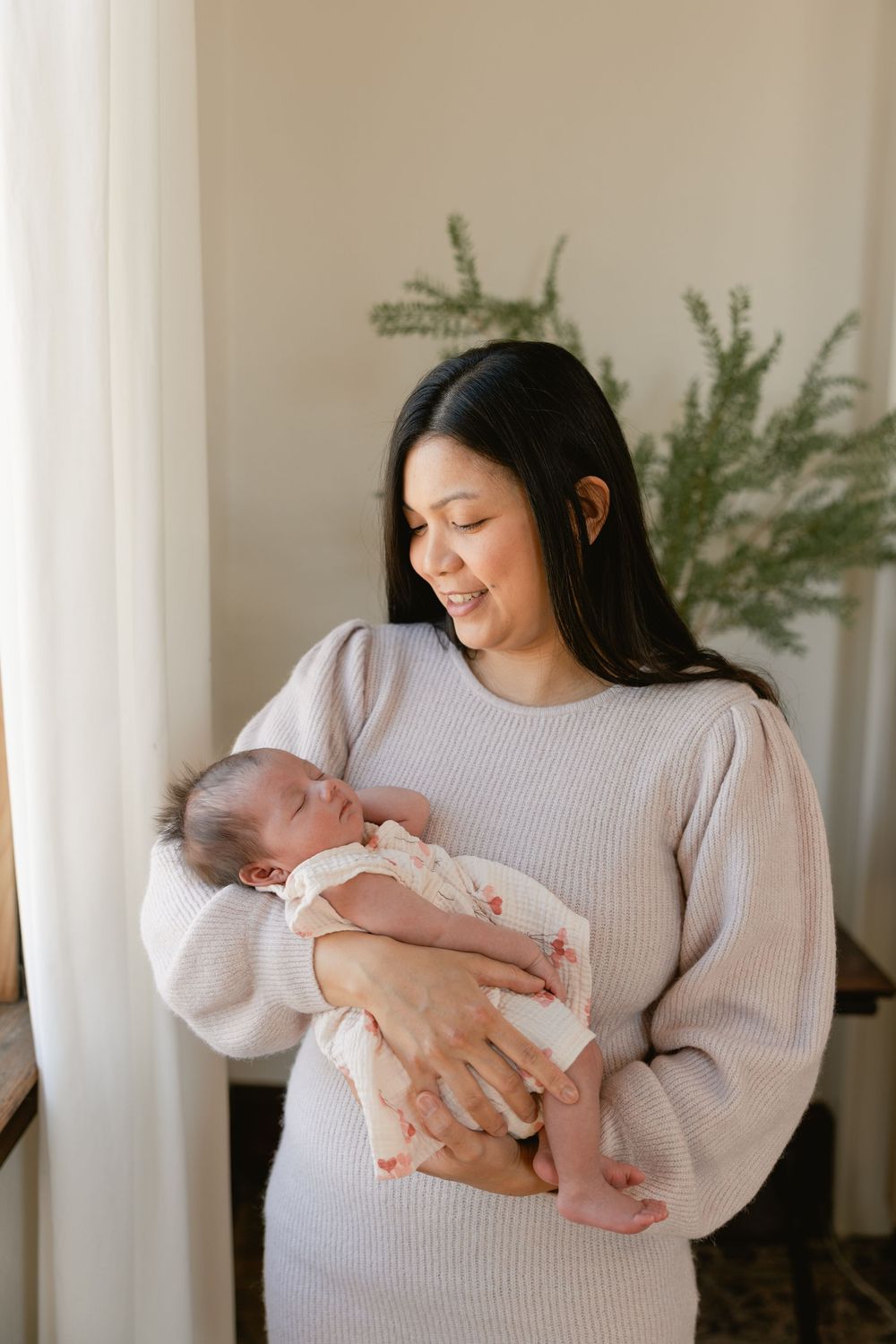 A person in a beige sweater cradles a sleeping newborn by a window with greenery.