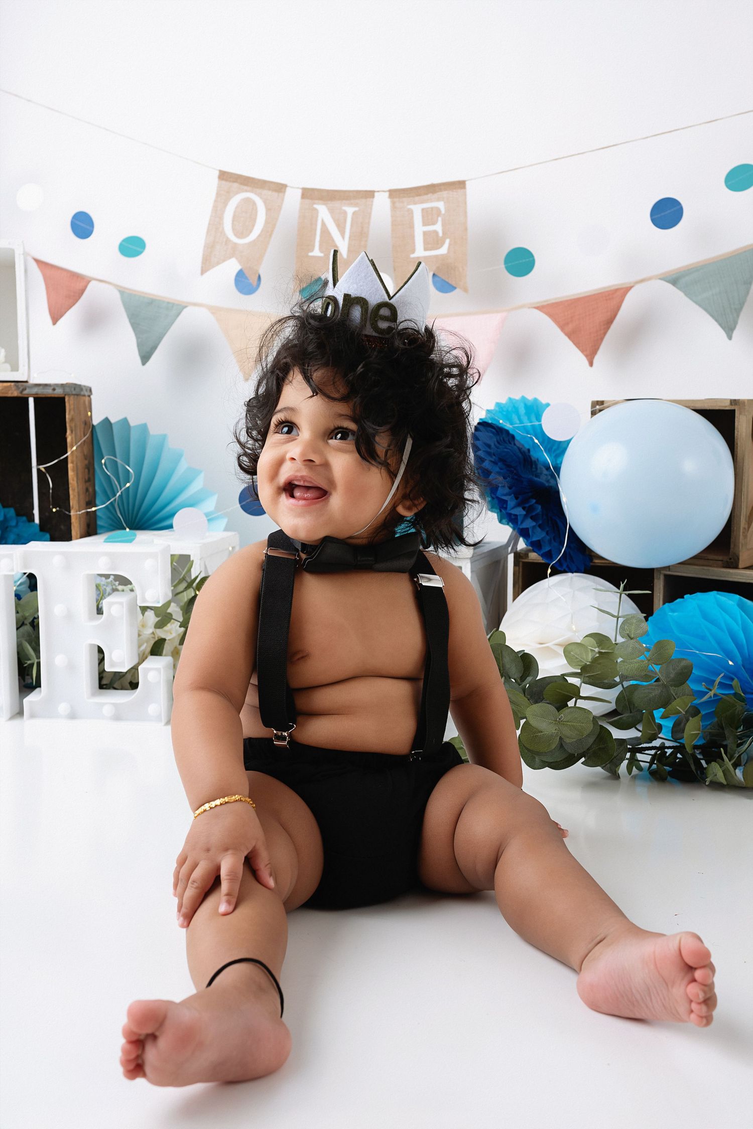 A baby in a black outfit lies on a white floor with birthday party decorations including balloons and banners in the background.