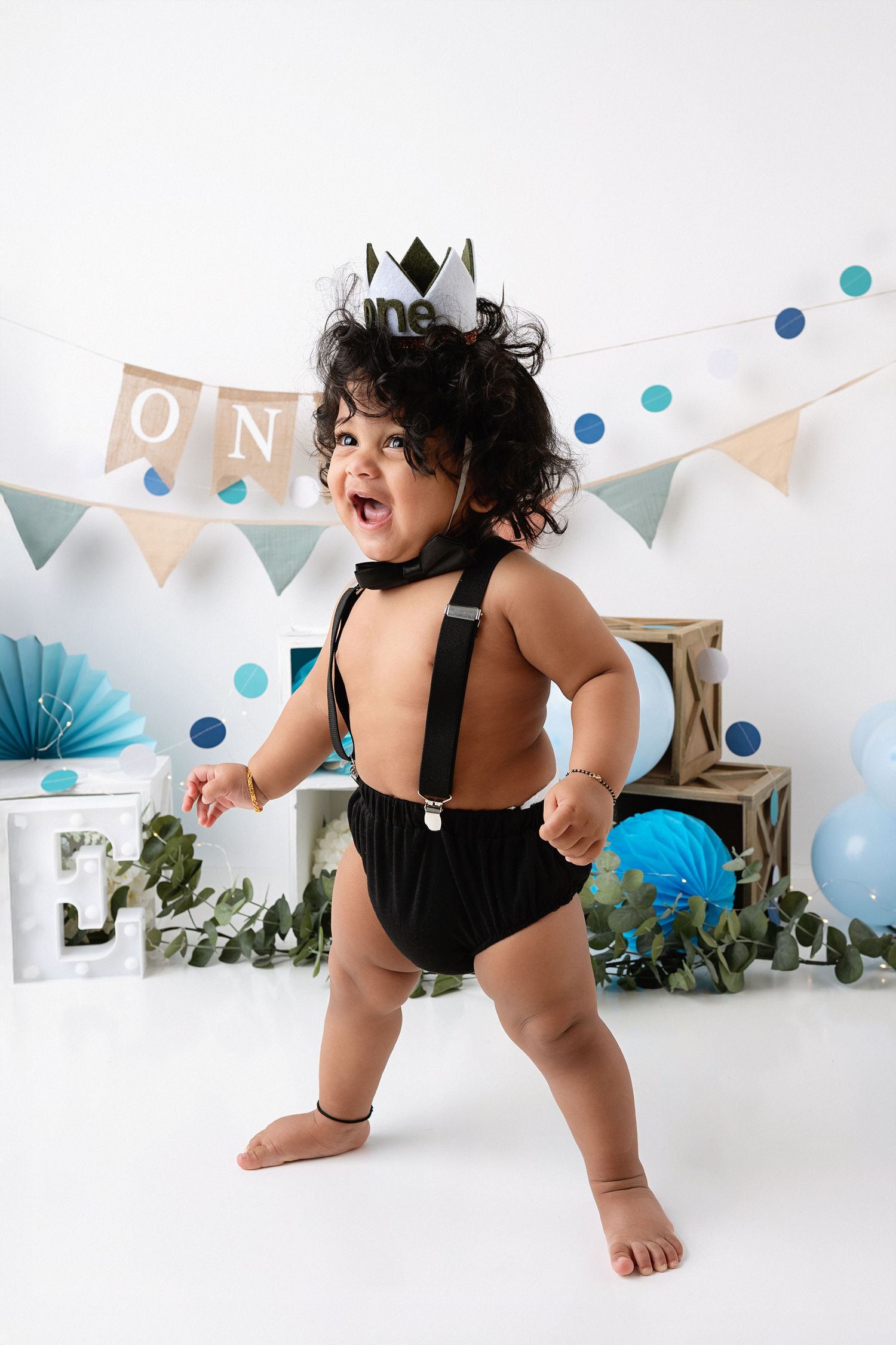 Baby in black swimsuit crawls on white floor with blue and white party decorations and banners in background.