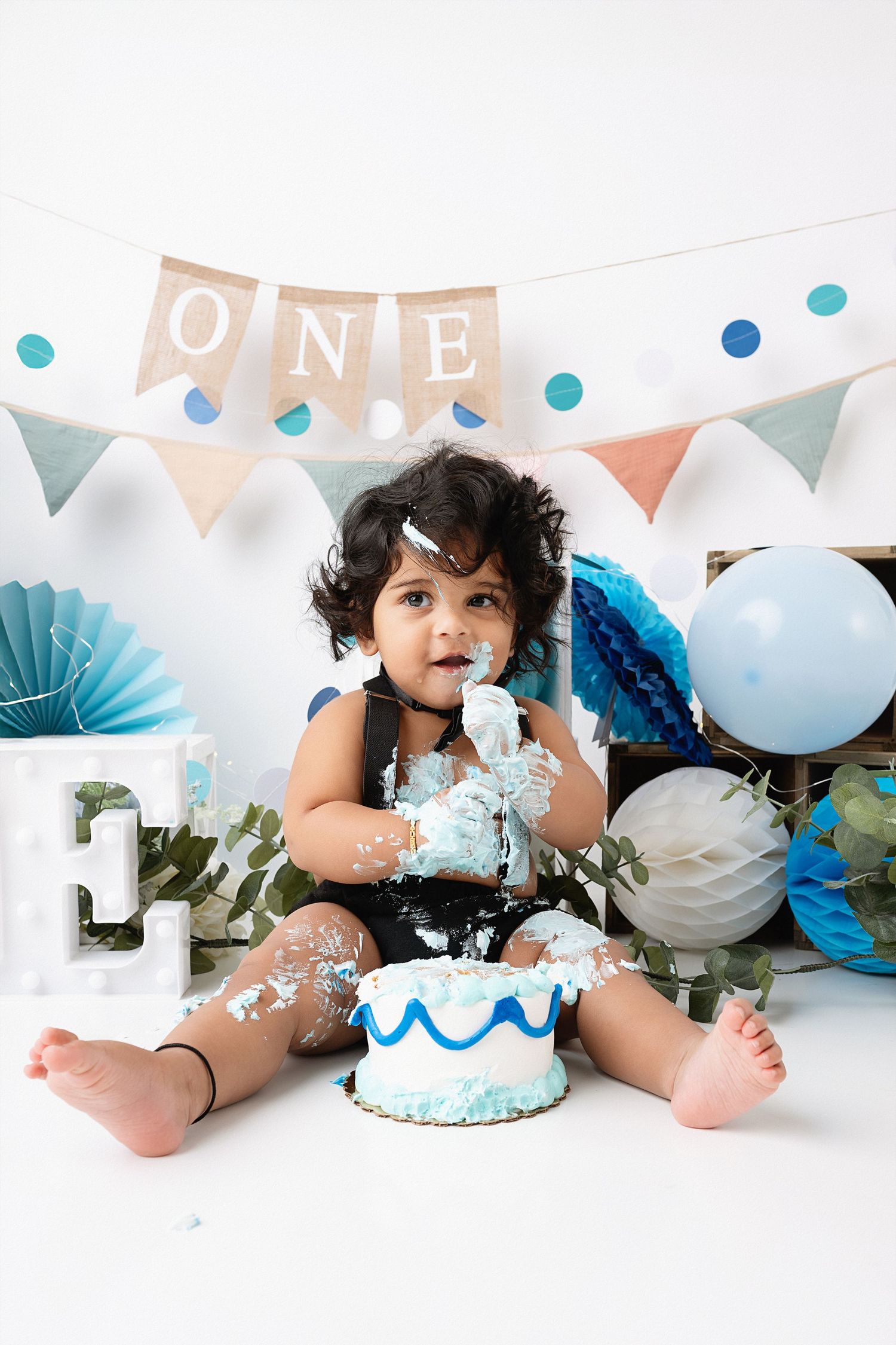 A baby in a sparkly outfit sits on a white backdrop with blue and white balloons and decorative banners for a cake smash.