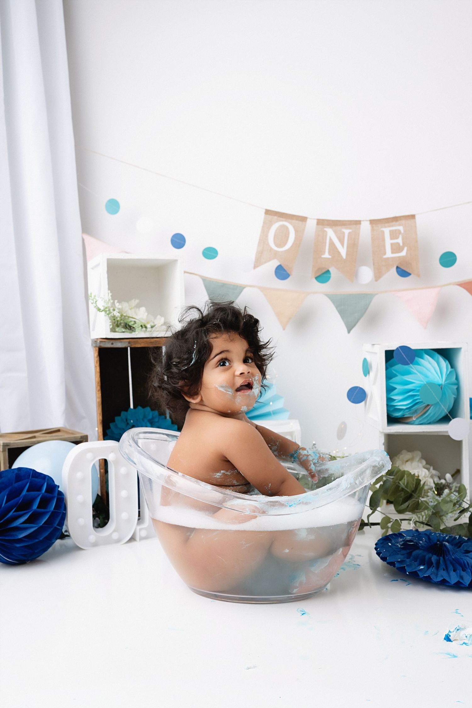 Baby's first birthday cake smash photo session with blue and white decorations and tissue paper pom poms.
