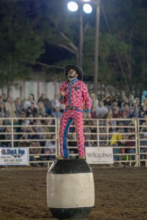 A rodeo clown in pink polka dot costume performs on barrel obstacles during nighttime show.