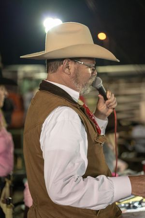 A cowboy wearing a brown vest and white shirt gestures while speaking at a nighttime event.