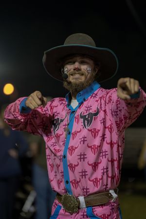 Cowboy wearing a pink patterned shirt with blue accents gestures during a nighttime rodeo event.