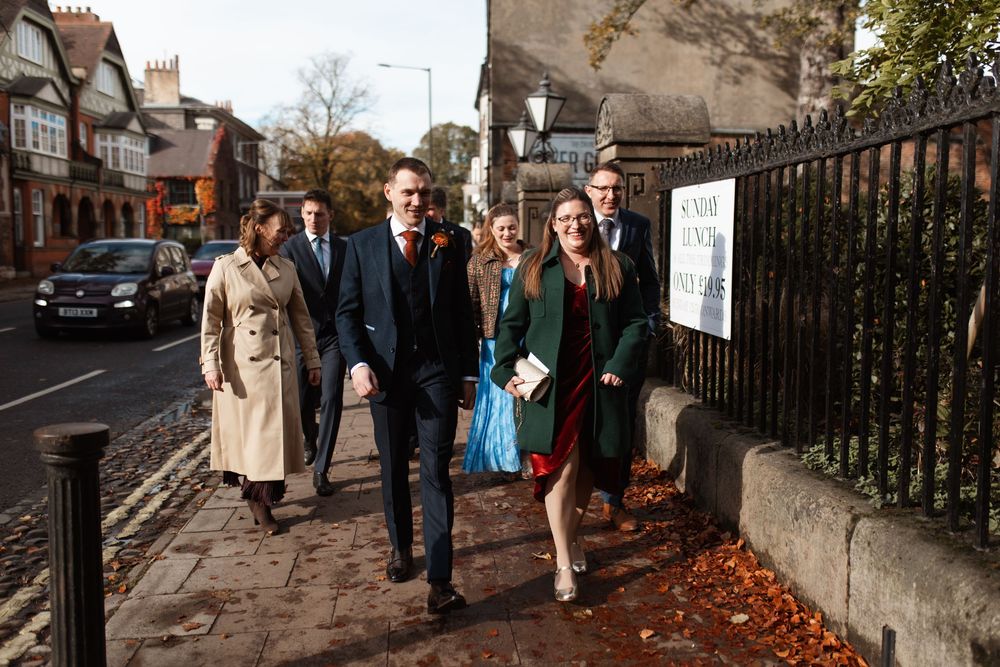 Group of people walking together on a leaf-covered sidewalk near a brick building on an autumn day.