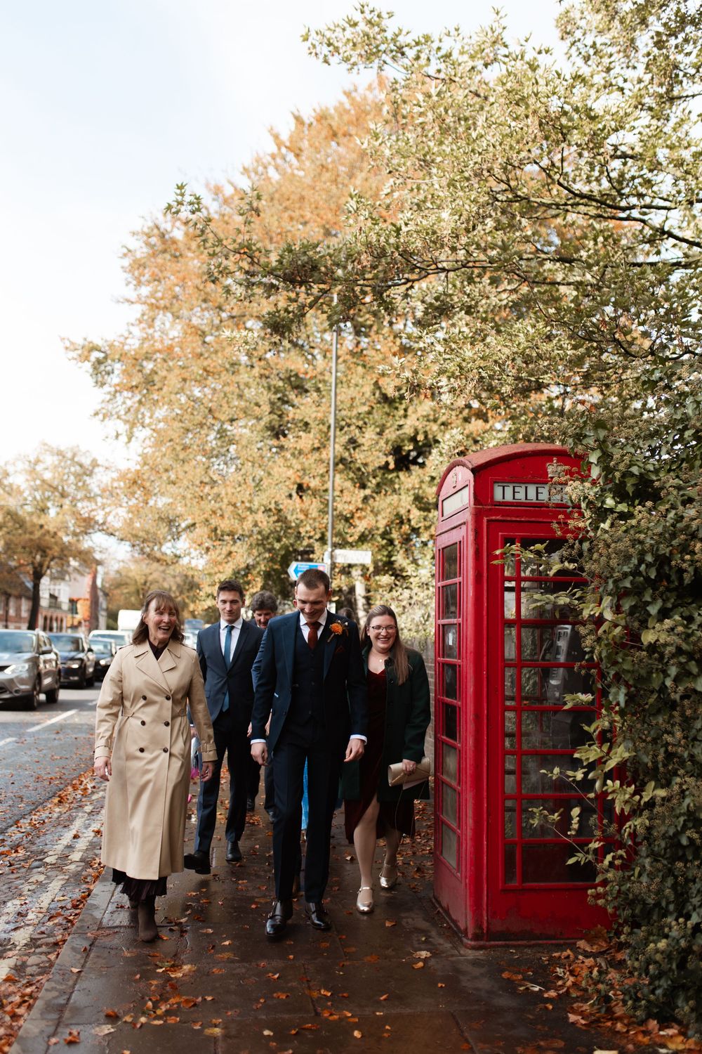 A group walks past a classic red British telephone booth on an autumn day with fallen leaves on the ground.