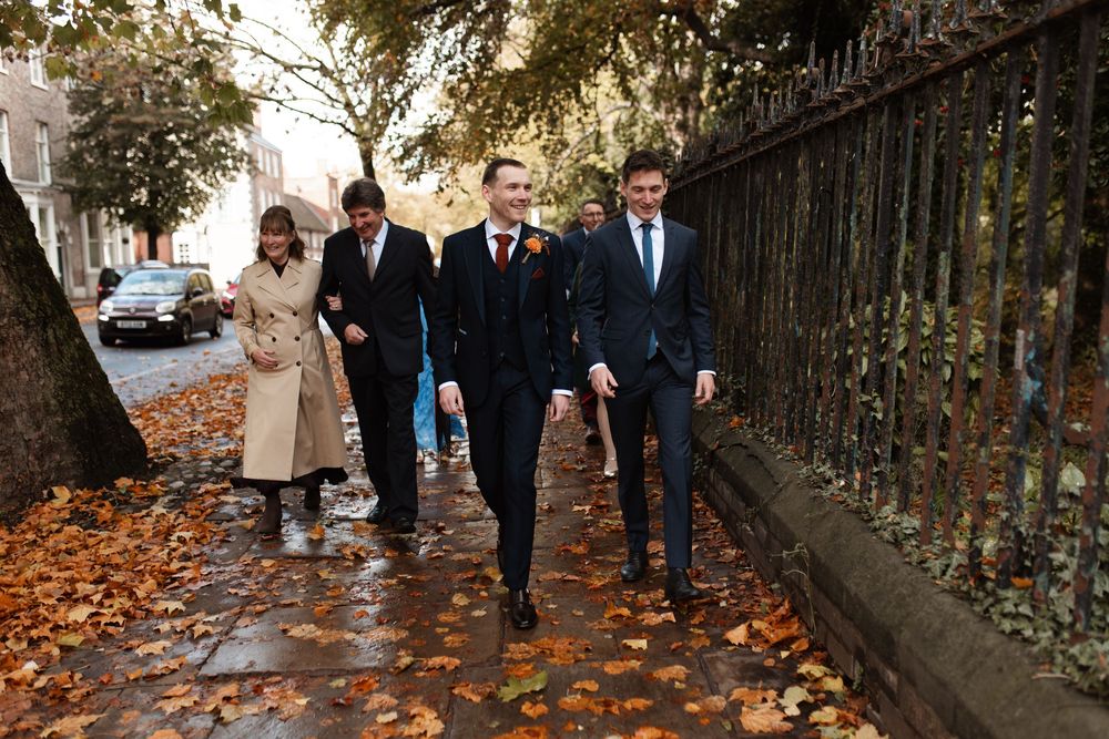 A wedding party walks along a leaf-covered path beside an iron fence on an autumn day.