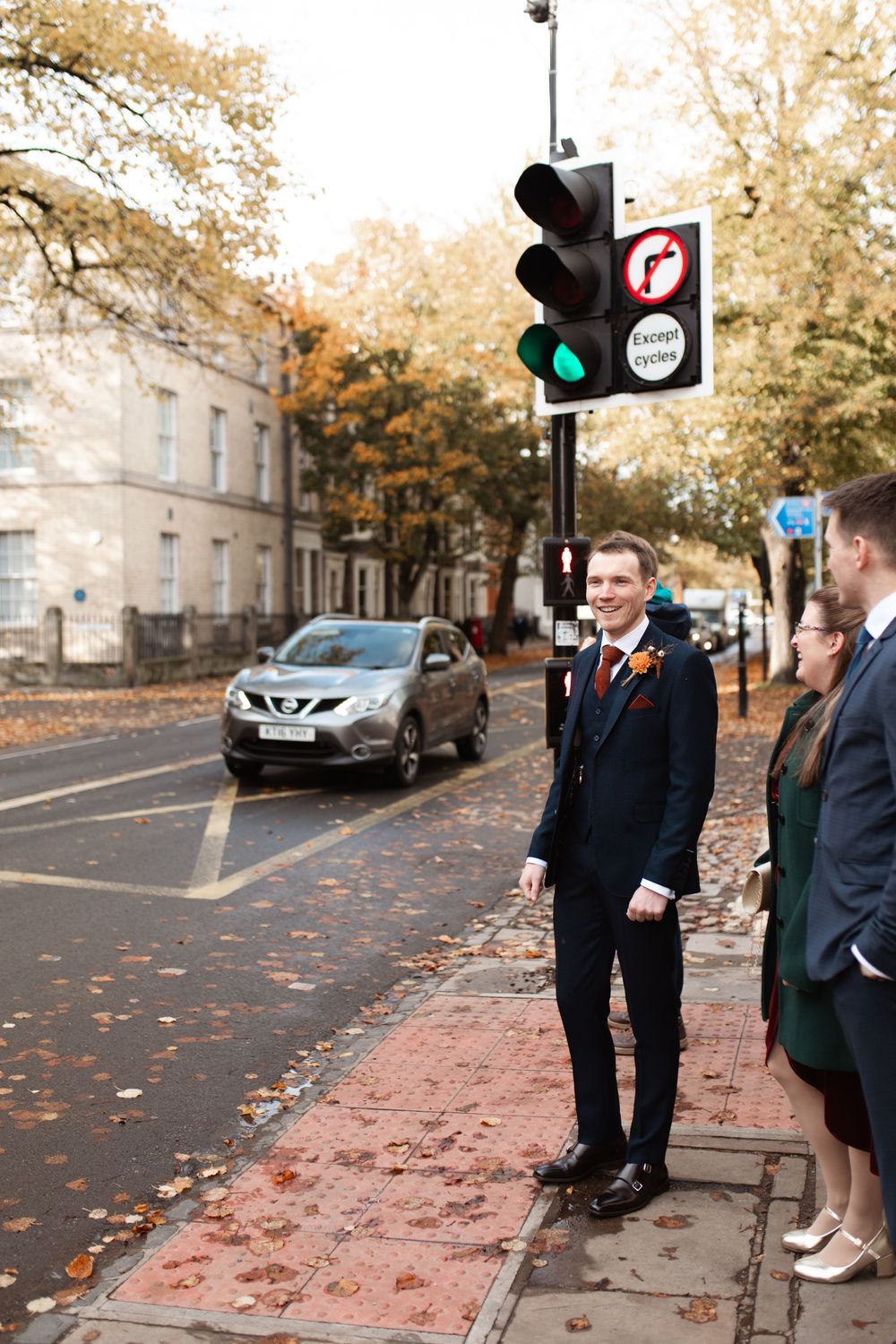 A group of formally dressed people walking down a brick sidewalk on an autumn day with fallen leaves.