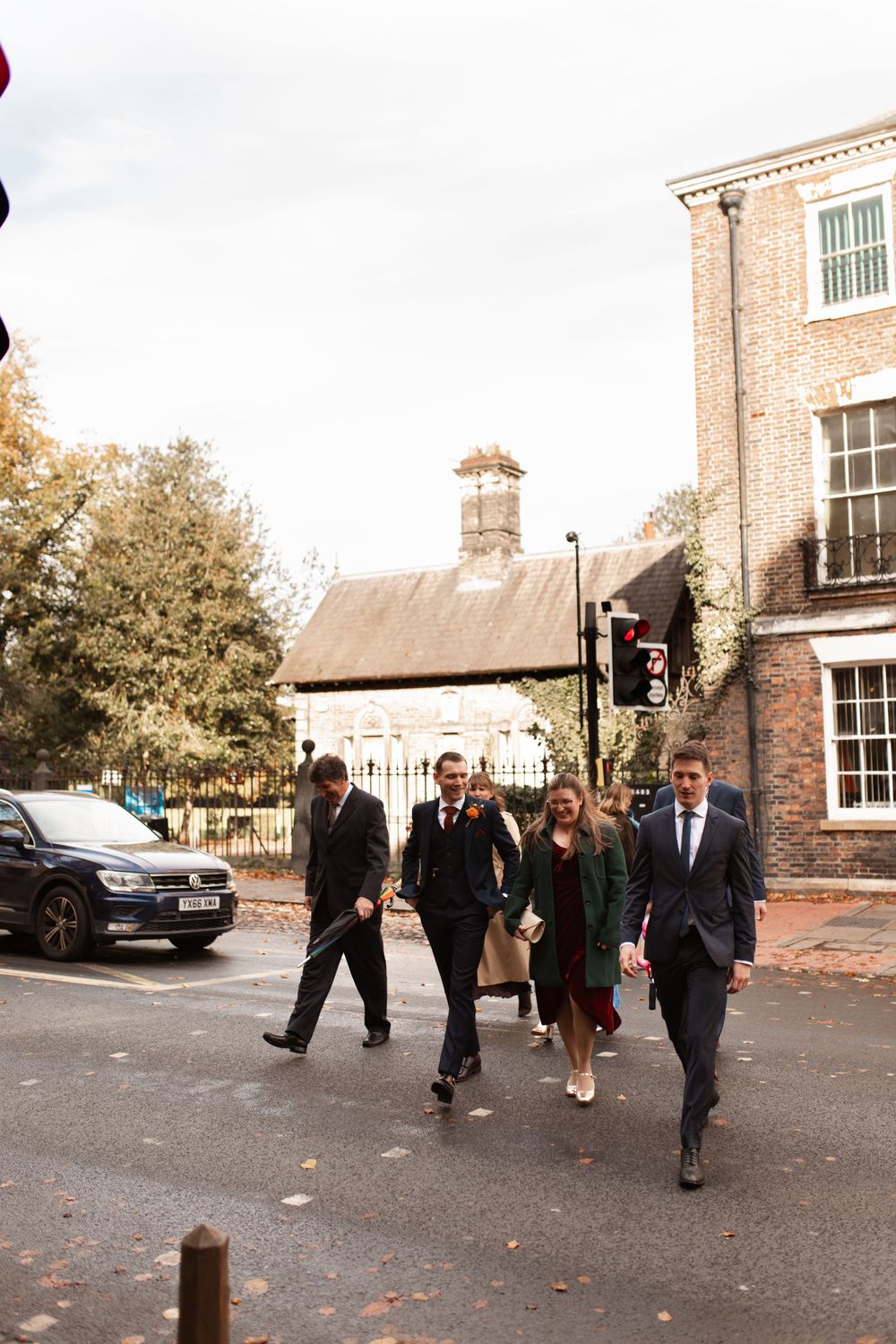 People in formal attire walking along a brick building exterior with fallen autumn leaves on the ground.