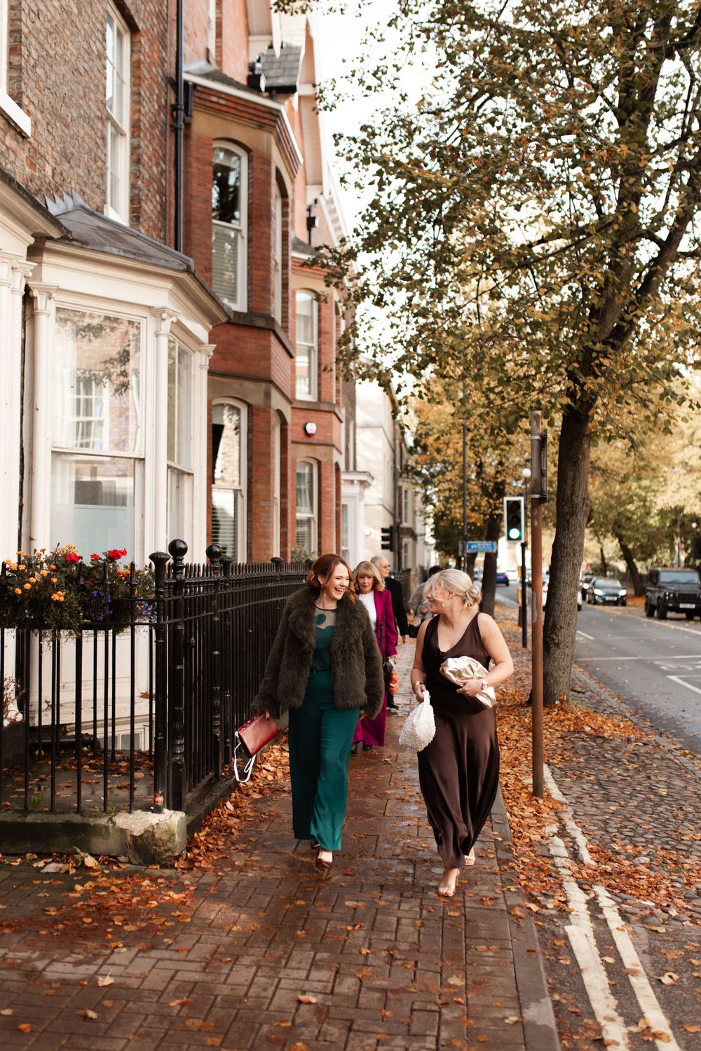 People stroll along a brick sidewalk beside historic brick buildings on a fall day with scattered leaves.