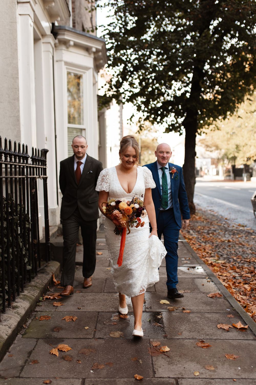 Wedding party walks along autumn sidewalk with scattered orange leaves on ground in urban setting.